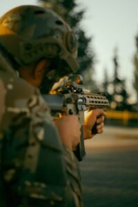 Back view of a soldier in uniform aiming a rifle outdoors during daytime exercise.